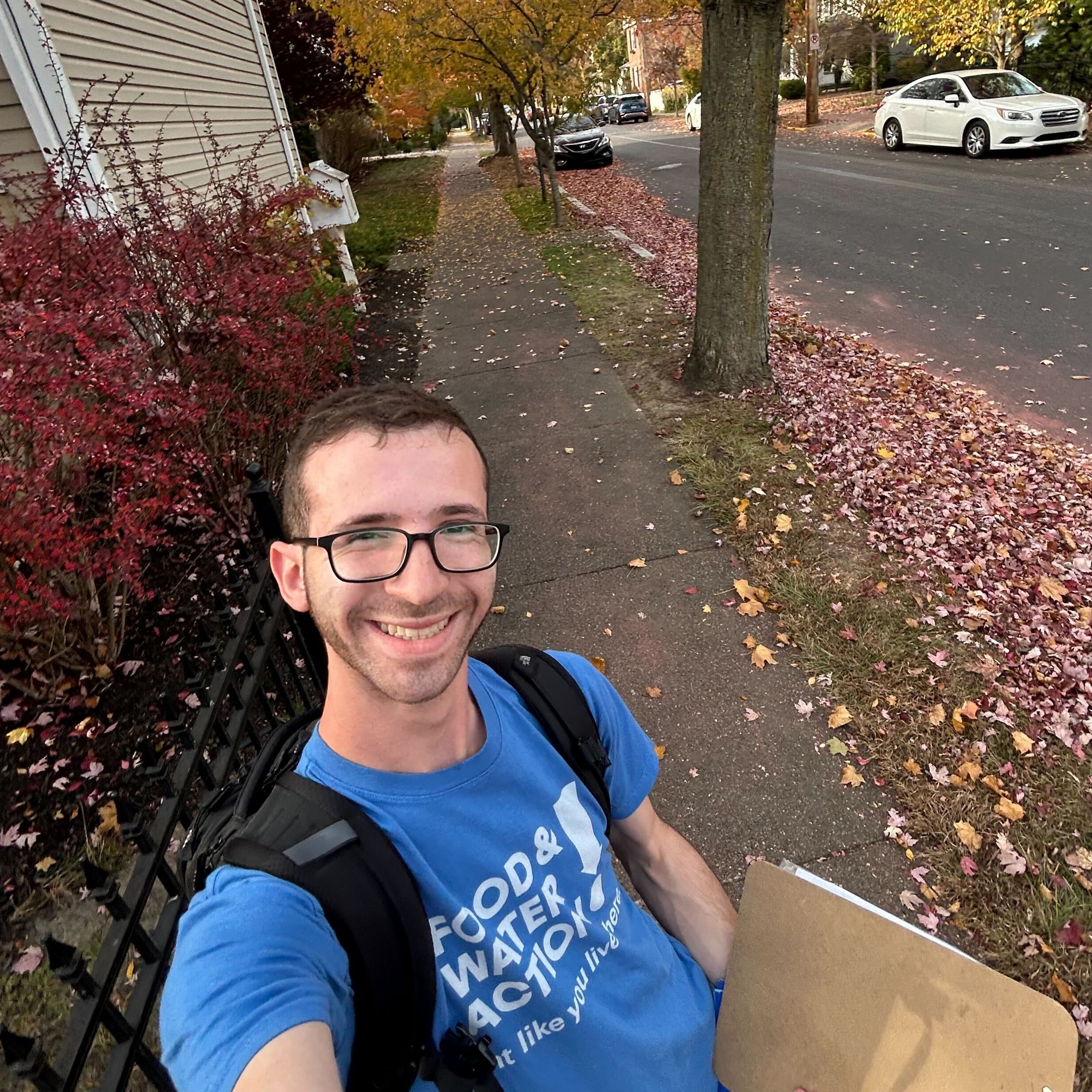 A white man holding a clip board stands on a shaded sidewalk, smiling at the camera.