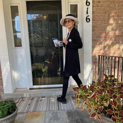 An older woman in a hat and sunglasses stands in front of a door, holding a stack of flyers