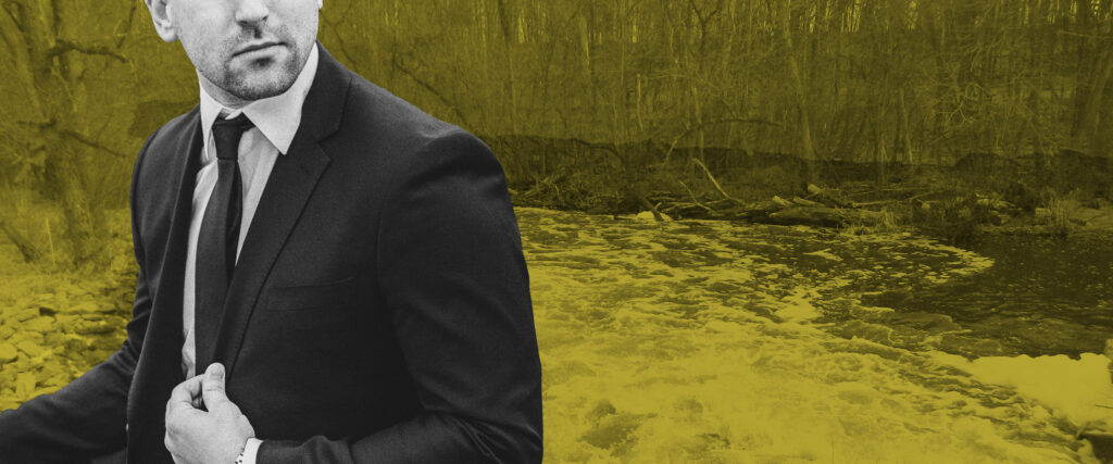 A man in a suit stands in front of a stream with a layer of PFAS pollution foam on the top.